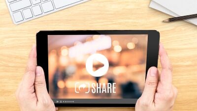 An overheard image of fair-skinned hands holding a smaller tablet device with a still image overalyed with a "play" icon and the word "SHARE" below the icon.