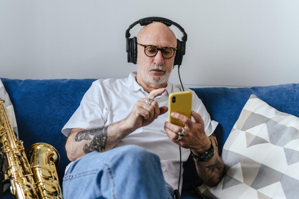 Elder Gen-Xer listening to a podcast on his iPhone using over-the-ear headphones on the couch with his saxophone propped up next to him.